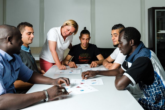 Lehrerin steht am Tisch und spricht mit fünf Menschen mit Migrationshintergrund