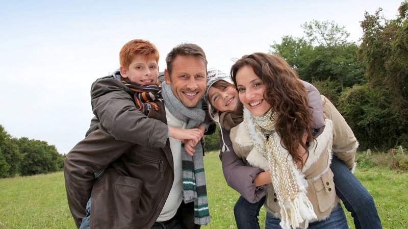 Eine vierköpfige Familie steht auf einer Wiese. Die Eltern tragen den Sohn und die Tochter huckepack.